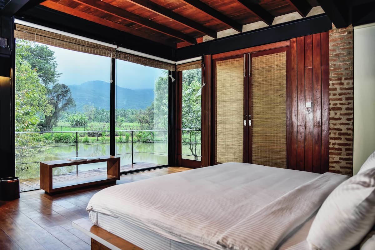 A bedroom with a large bed, wooden flooring, a bench, and a view of greenery and mountains through large windows.