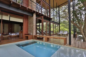 Un área de piscina moderna al aire libre con mobiliario de terraza de madera y un nivel superior con cerramiento de vidrio.