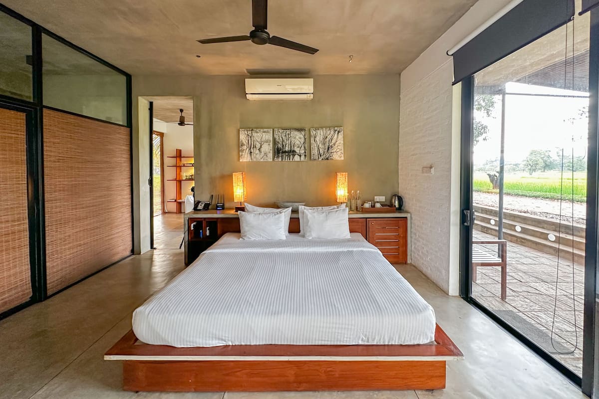 A bedroom with a large bed, ceiling fan, artwork on the wall, and a view of a garden through a large window.