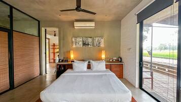 A bedroom with a large bed, ceiling fan, artwork on the wall, and a view of a garden through a large window.