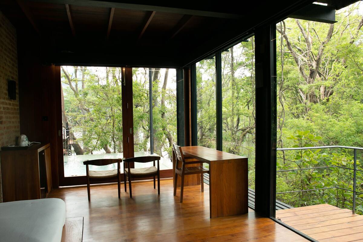A room with a wooden floor, a desk, two chairs, and large windows offering a view of a forest.