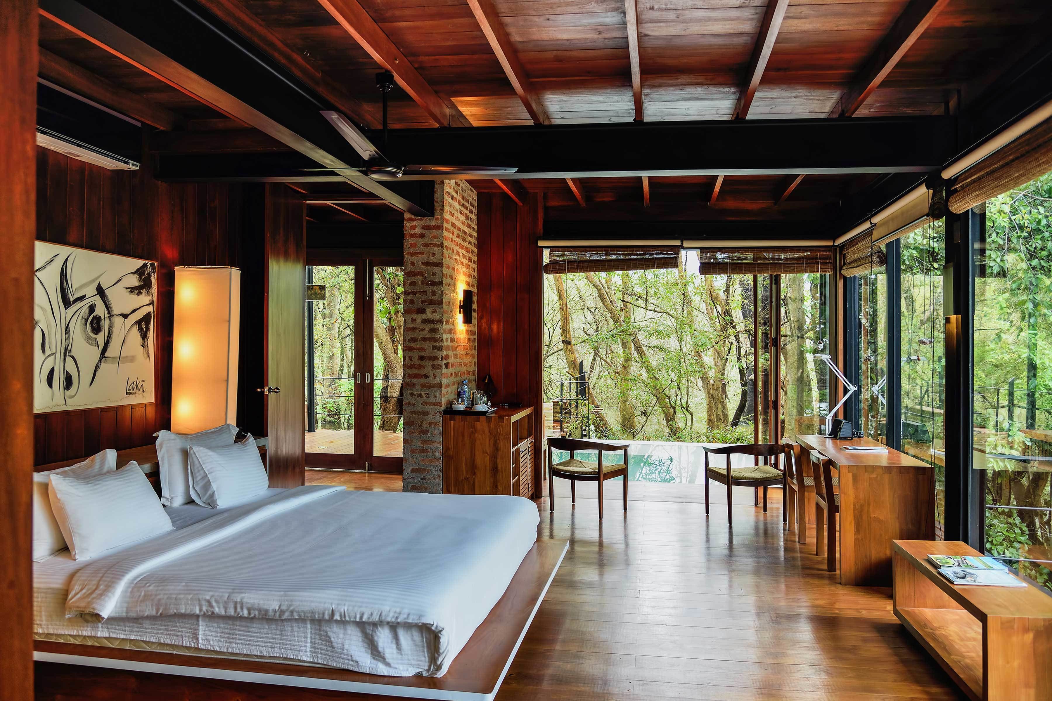 A spacious bedroom with a large bed, wooden furniture, and a view of the forest through large windows.