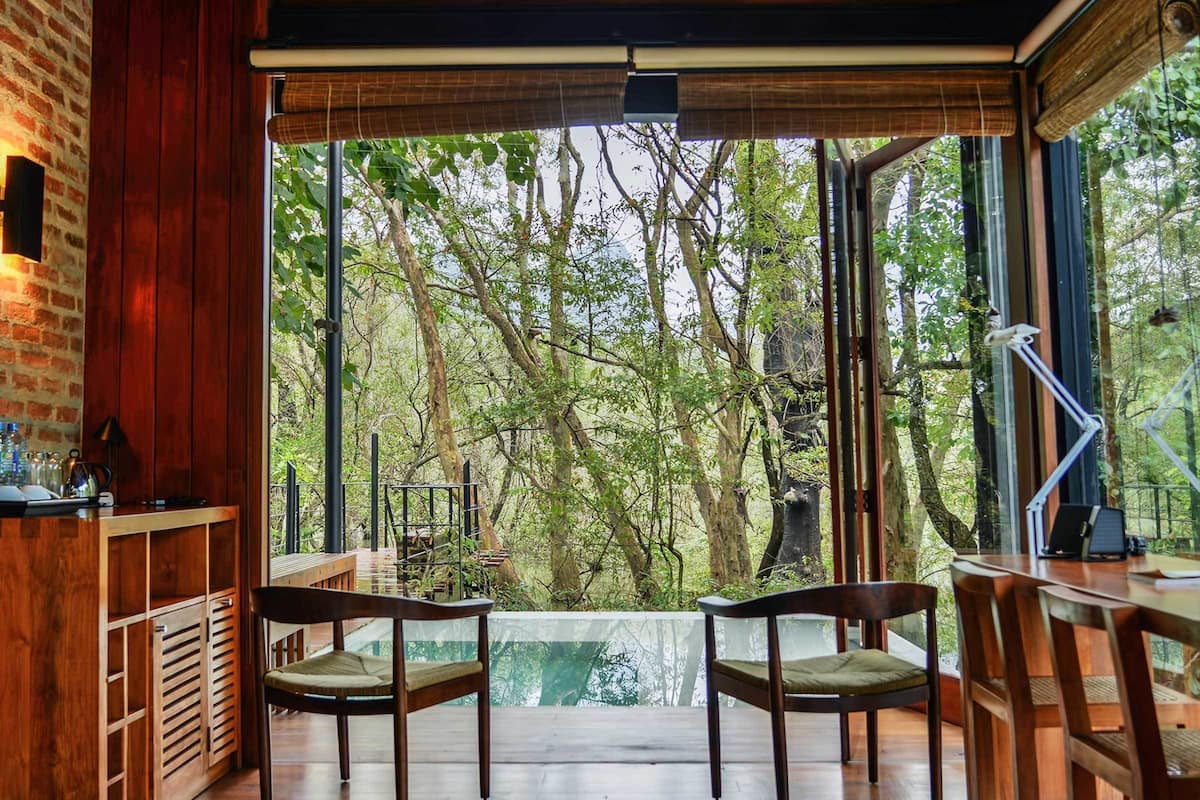 A spacious room with wooden flooring, a brick wall, and a large window offering a view of trees.