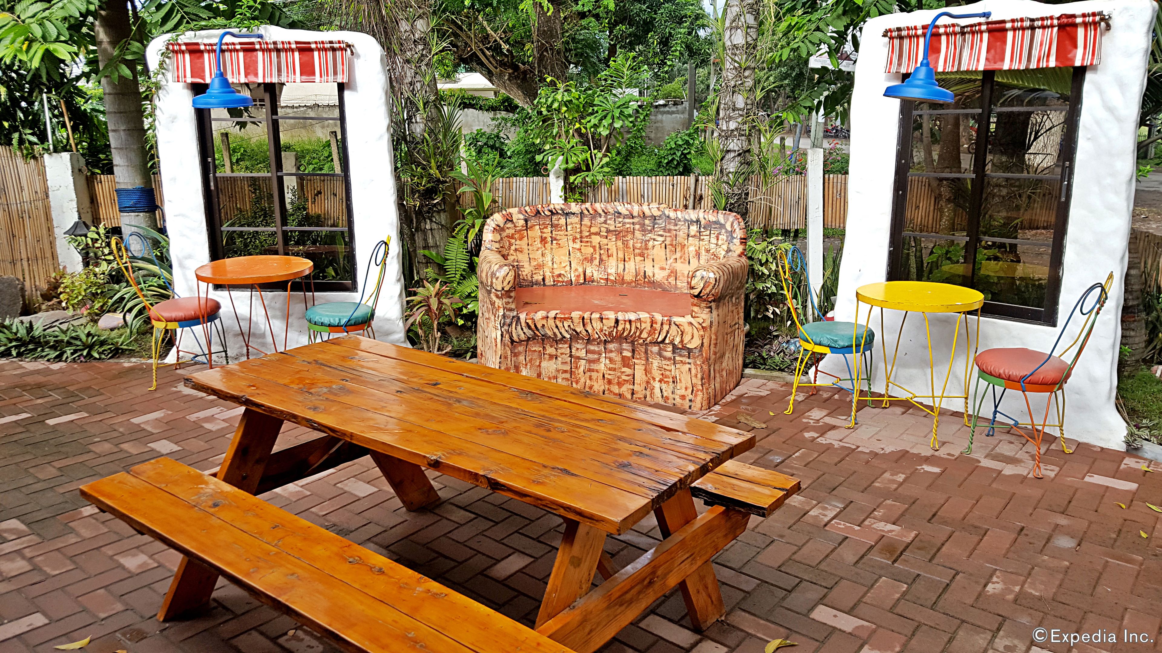 A wooden picnic table with benches, surrounded by colorful chairs and tables, set in a garden with a white wall and windows.