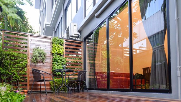 Un patio moderno al aire libre con una terraza de madera, sillas de metal y un ventanal que da a un espacio interior.