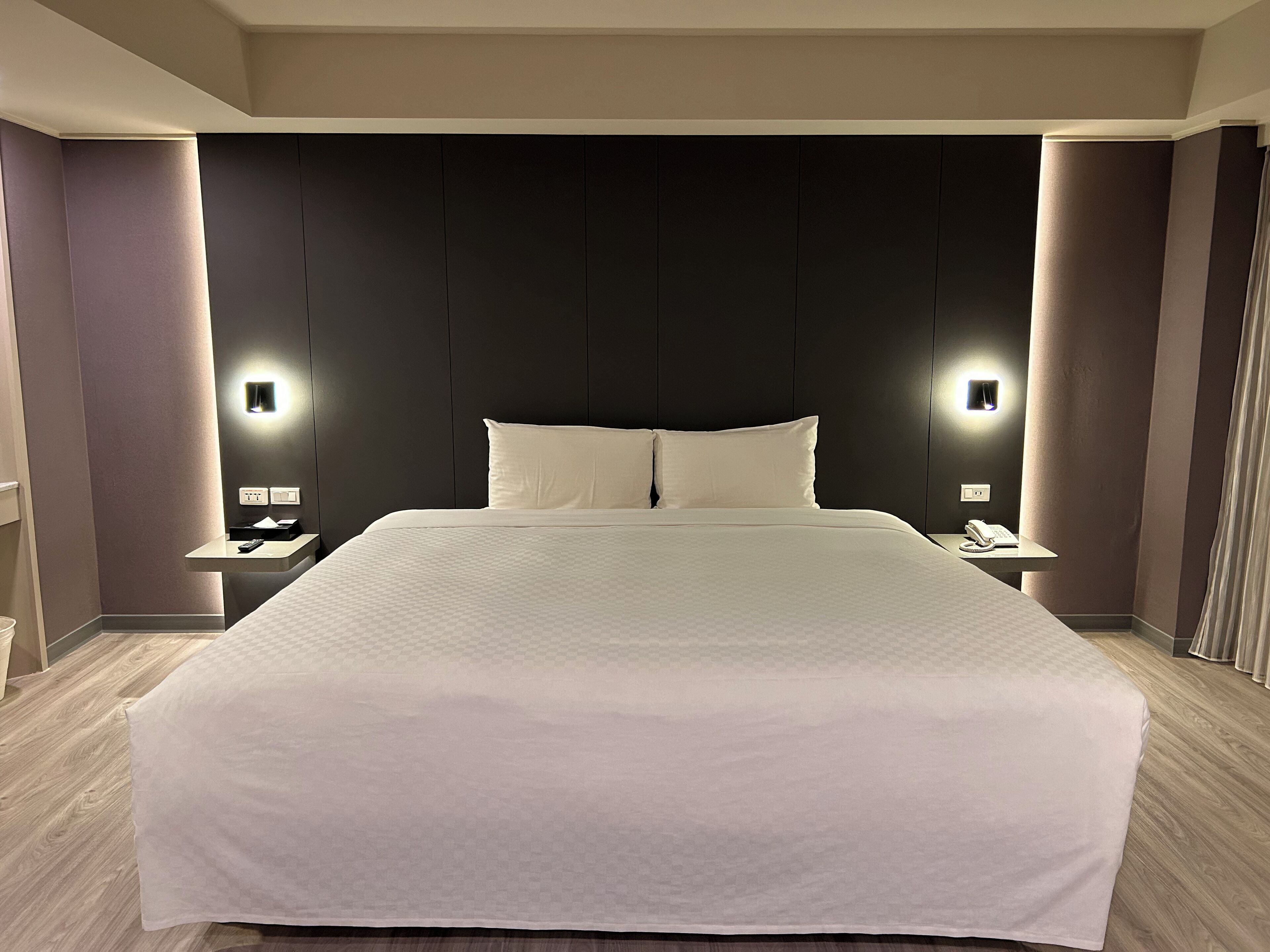 A modern hotel room with a large bed, two bedside tables with lamps, and a dark headboard.