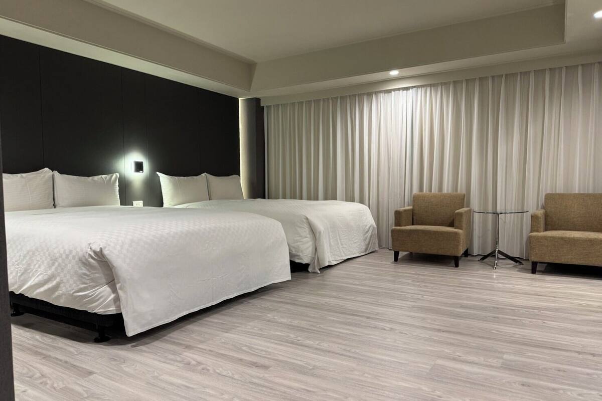 A modern hotel room with a large bed, two bedside tables, a seating area with two armchairs, and a large window with curtains.