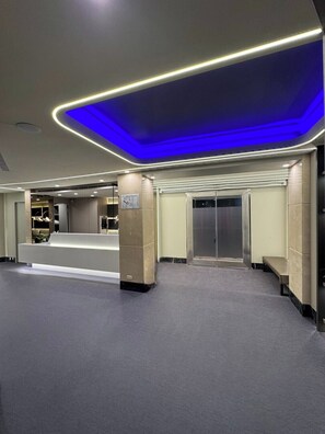 Un hall amplio con una recepción, un ascensor de vidrio y un techo iluminado en azul.