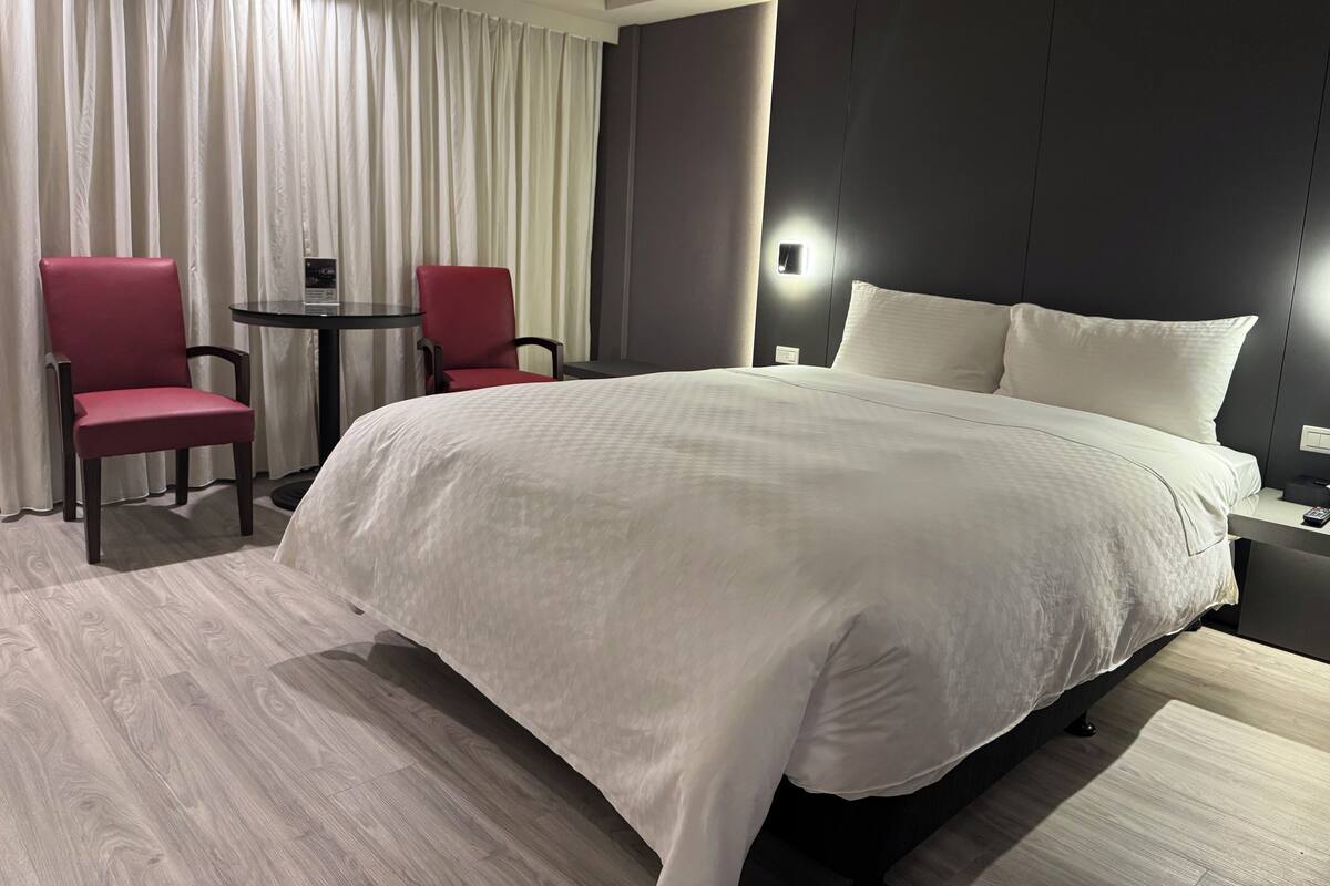 A hotel room with a large bed, two red chairs, a small table, and a wall-mounted light.