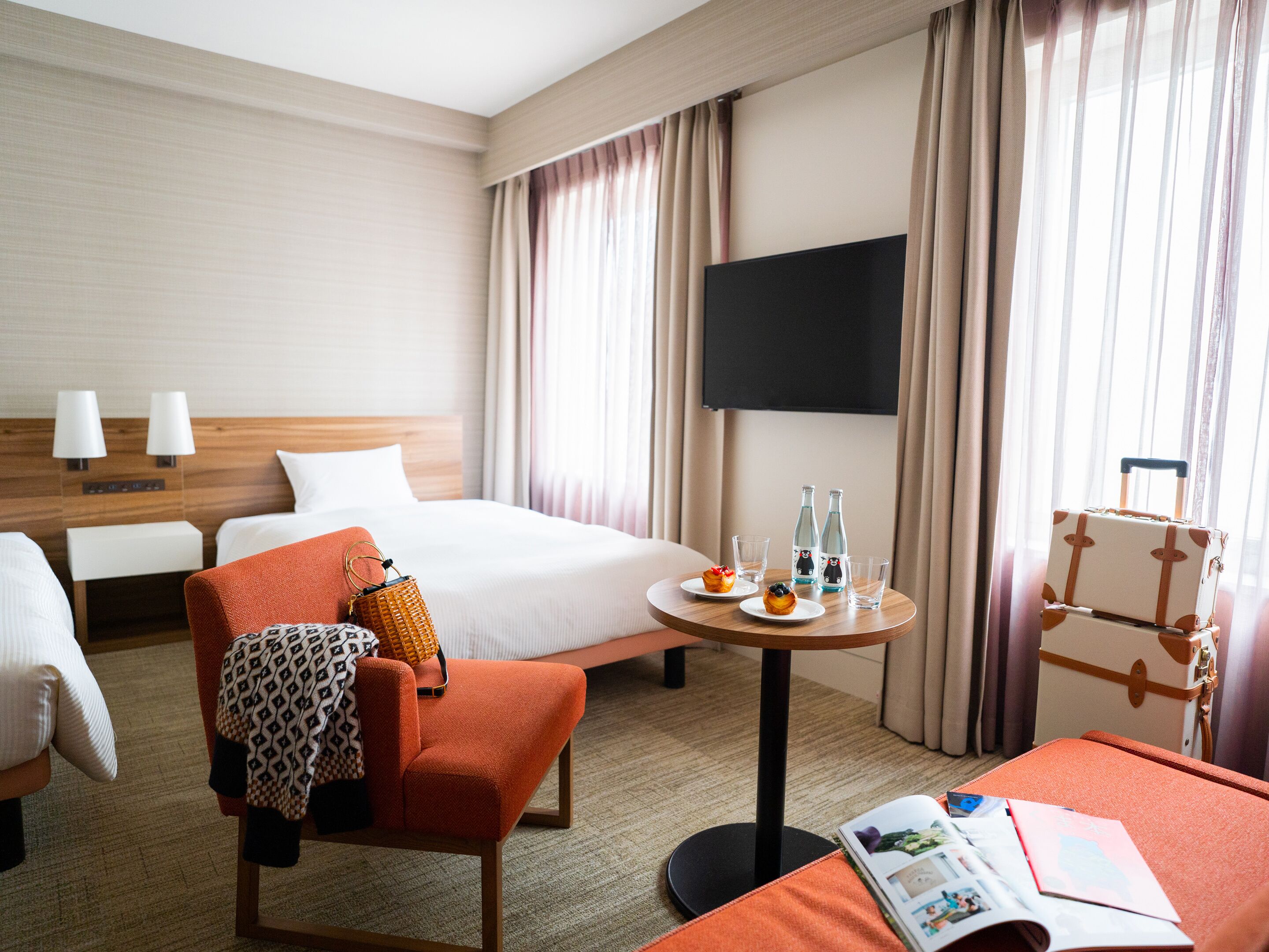 Habitación de hotel con cama, una silla, una mesita con bebidas y una revista, televisión y maleta.