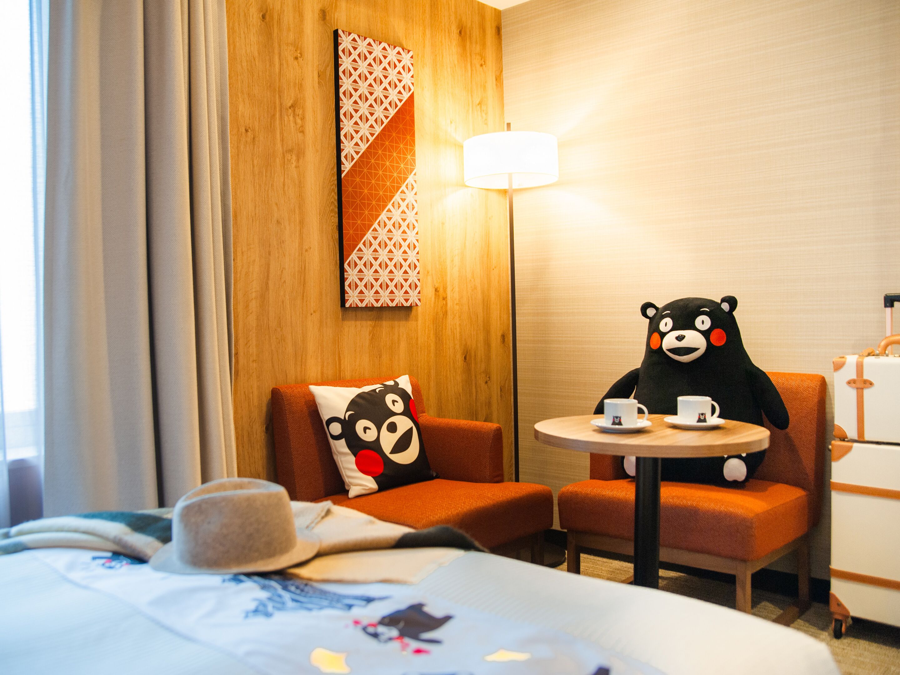 Habitación de hotel con una cama, un oso de peluche, dos sillas, una lámpara y una pared con diseño geométrico.