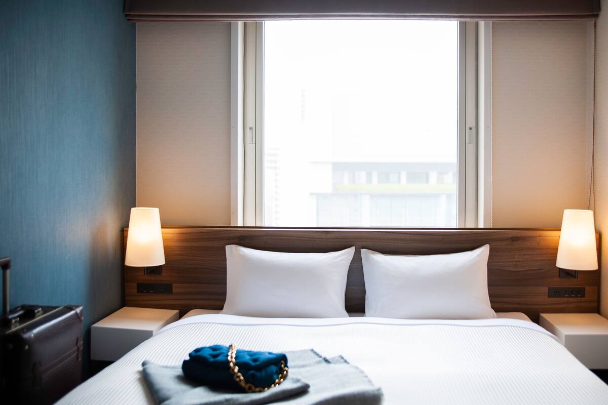 Een hotelkamer met een groot bed, twee bedlampjes, een raam met zonwering en uitzicht op een gebouw.