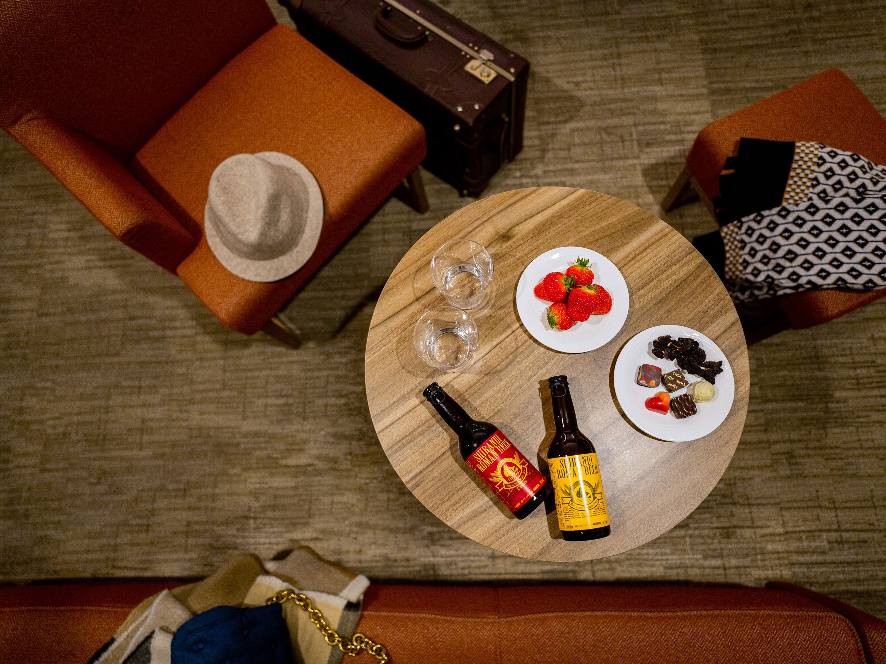 A round wooden table with a bottle of beer, a bottle of wine, a plate with strawberries, and a plate with chocolate-covered strawberries.