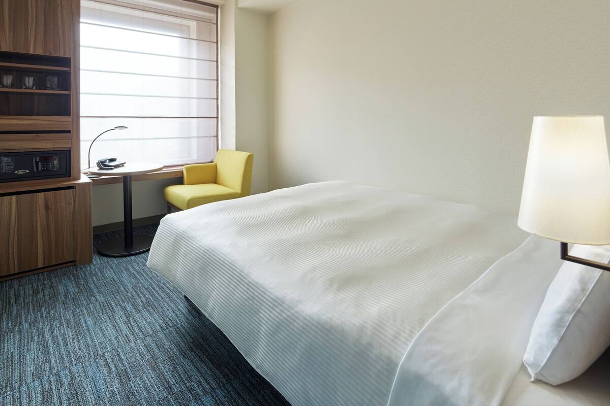 Hotelkamer met een bed, een bureau met stoel, een lamp en een raam met jaloezieën.