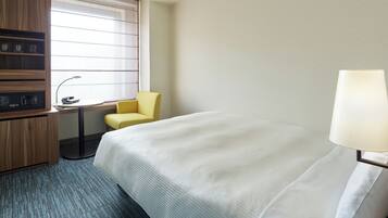 Una habitación de hotel con cama, un escritorio con silla, una lámpara y una ventana con persianas.