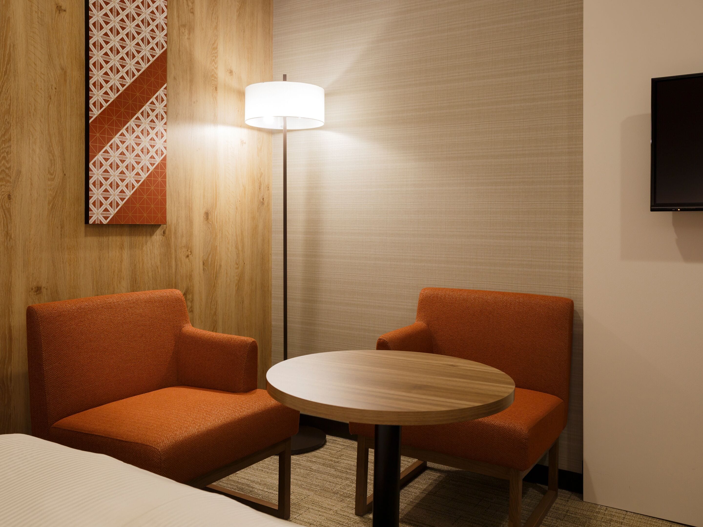A hotel room with two orange armchairs, a round wooden table, a floor lamp, a TV mounted on the wall, and a bed with a white headboard.