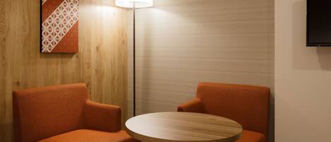 A hotel room with two orange armchairs, a round wooden table, a floor lamp, a TV mounted on the wall, and a bed with a white headboard.