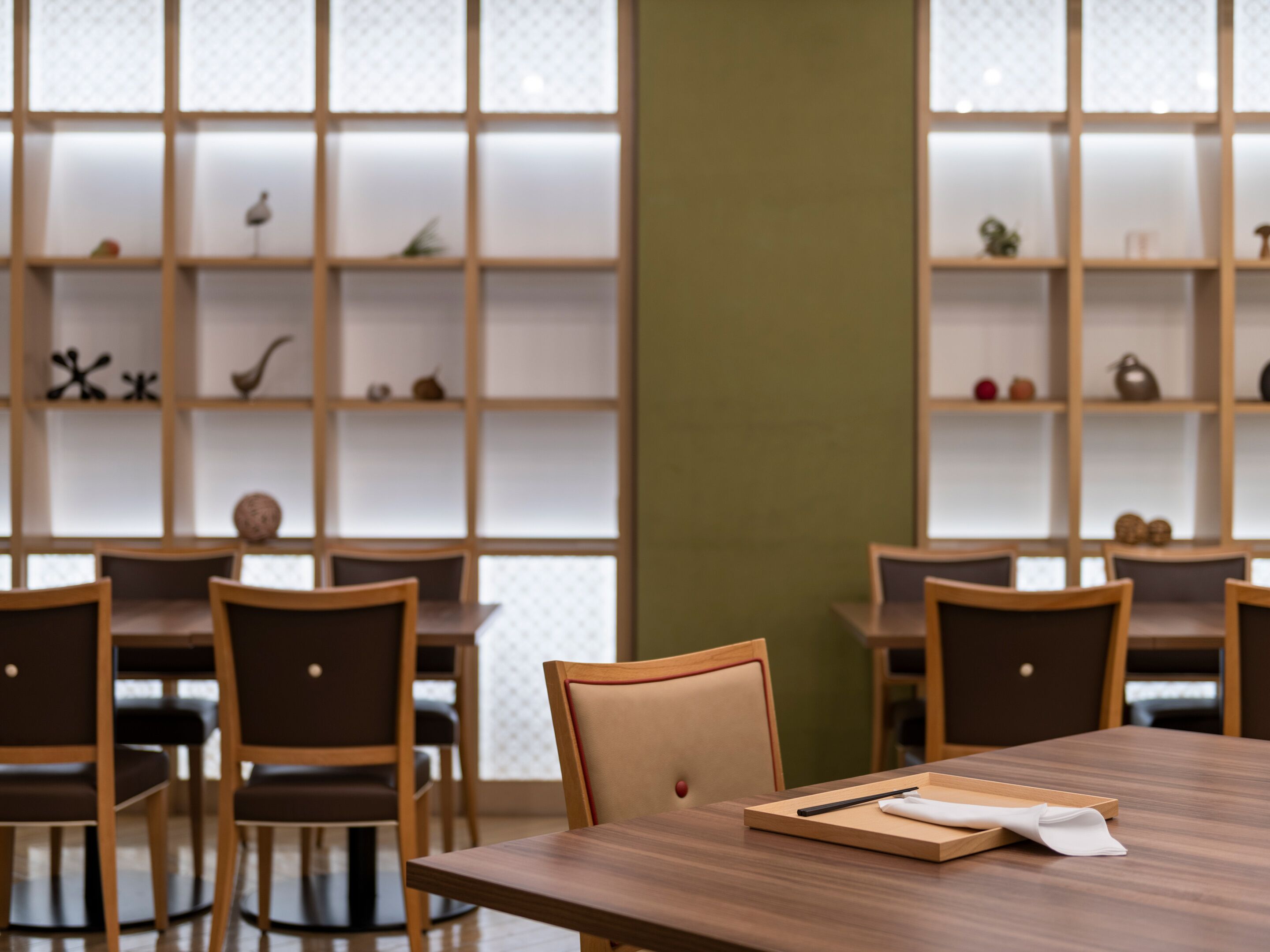A modern dining area with wooden tables and chairs, a large window with frosted glass panels, and shelves with decorative items.