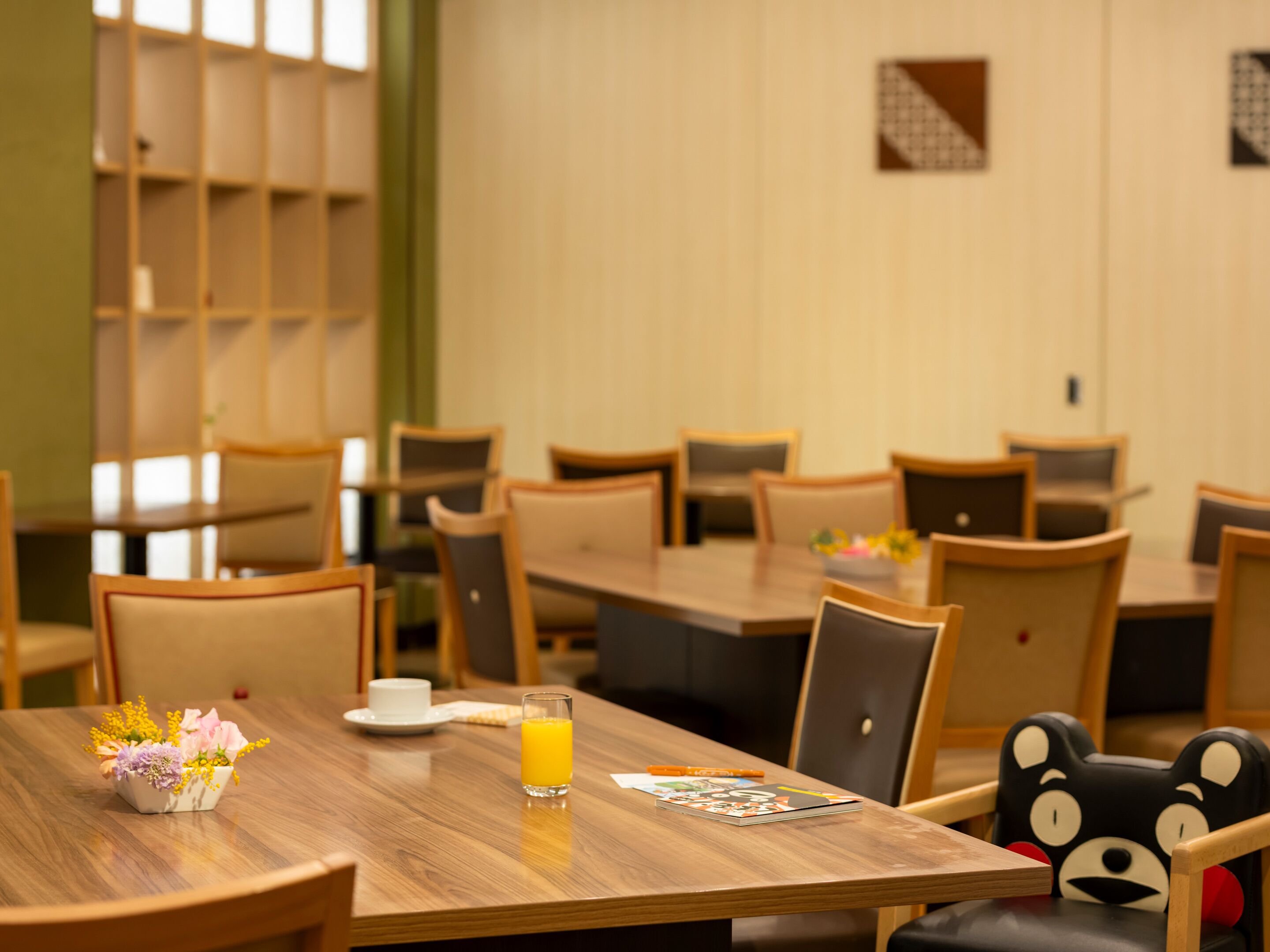 Un coin repas avec des tables et des chaises en bois, un petit vase avec des fleurs, un verre de jus d’orange et un livre posé sur la table.