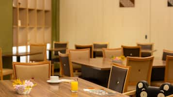Un coin repas avec des tables et des chaises en bois, un petit vase avec des fleurs, un verre de jus d’orange et un livre posé sur la table.