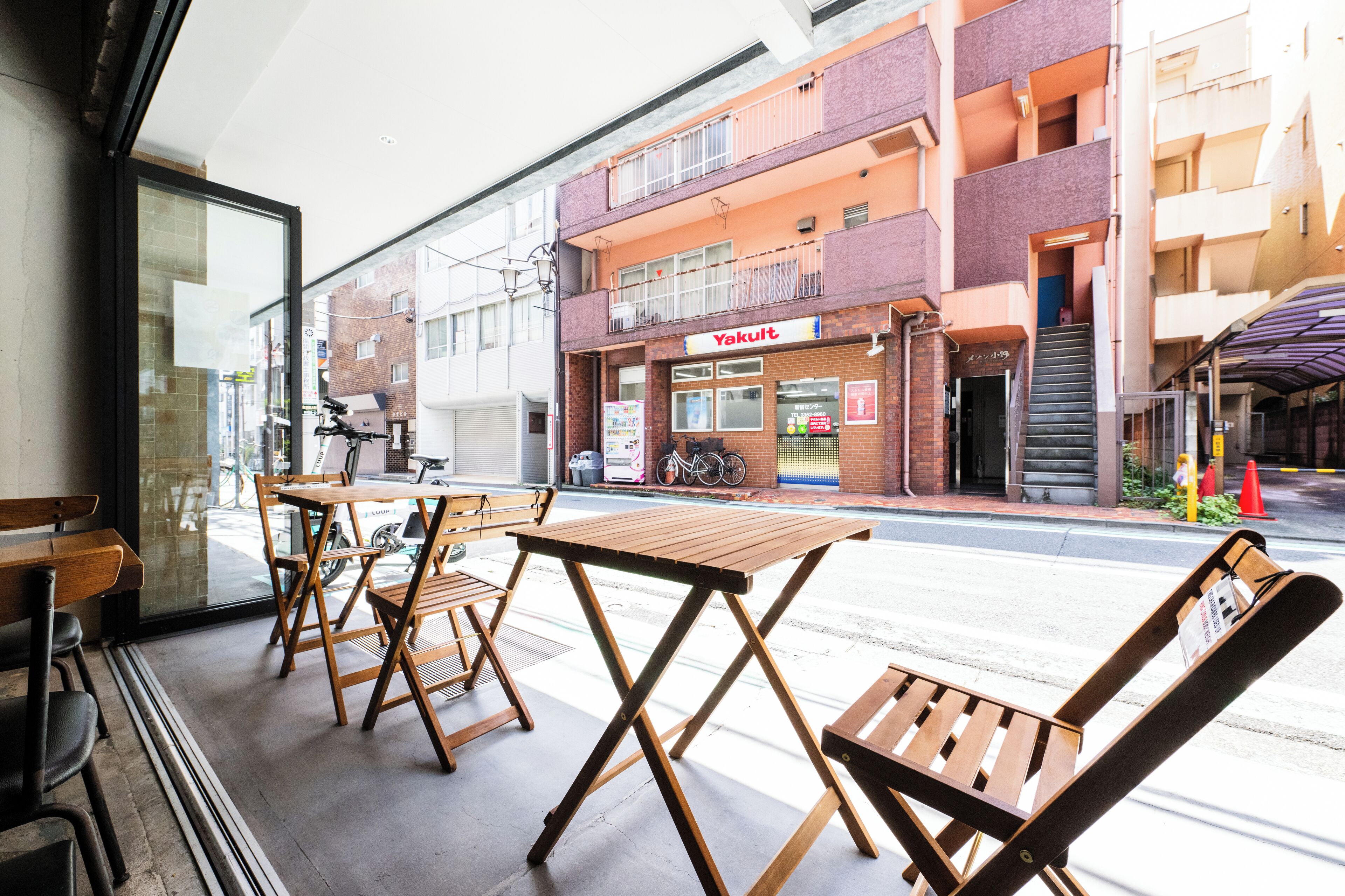 A street view with a restaurant named 'Yakult' on the corner, outdoor seating with wooden tables and chairs, and a bicycle parked outside.