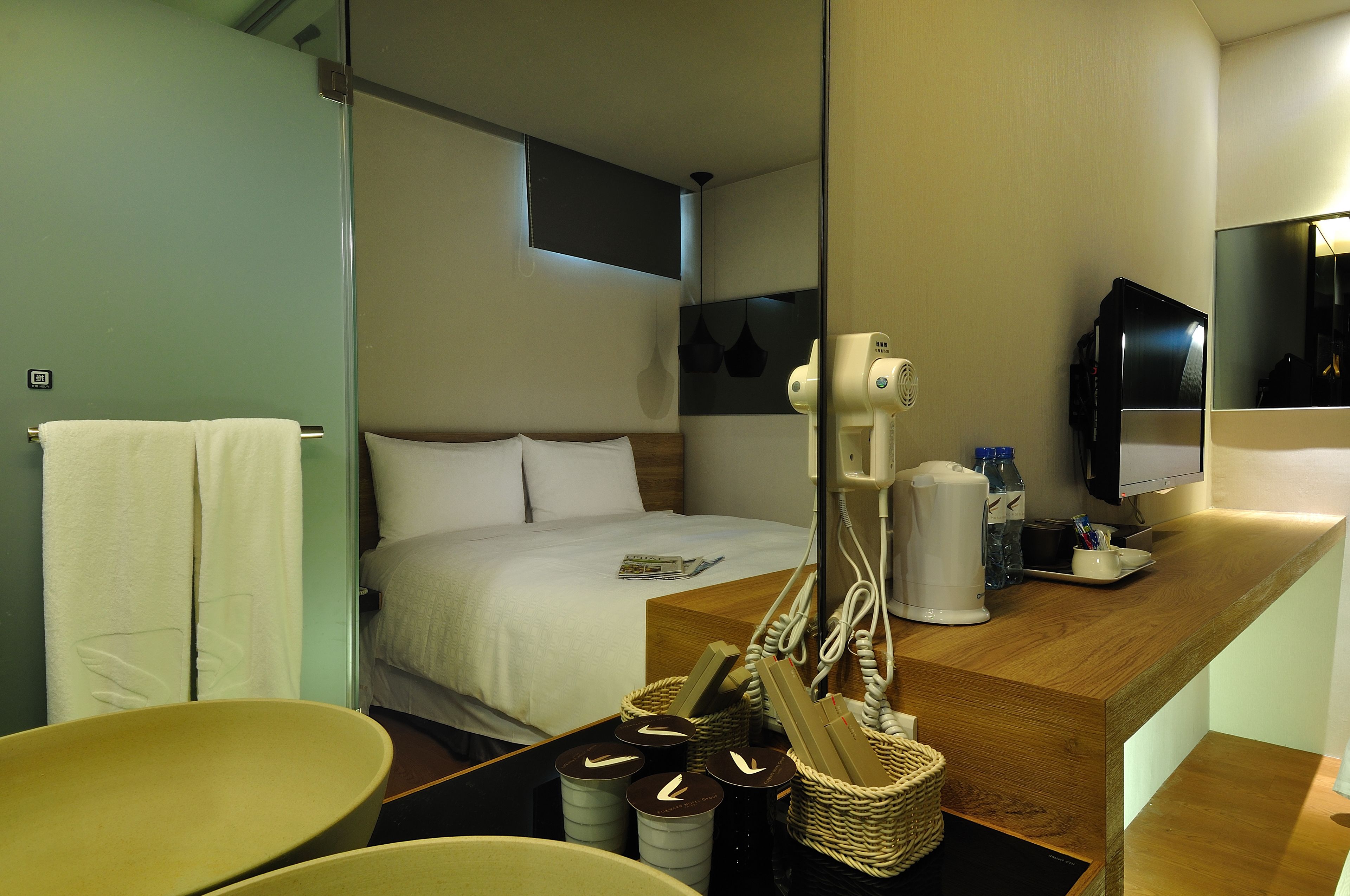 Ein Hotelzimmer mit einem Bett, einem Holztisch, einem Spiegel, einem Fernseher und einem Badebereich mit Handtuchhalter und Waschbecken.