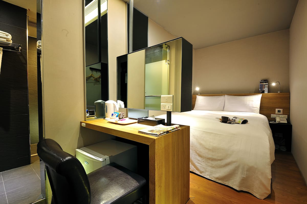 Una habitación de hotel con cama, escritorio con silla, espejo y mesita de noche con lámpara y teléfono.