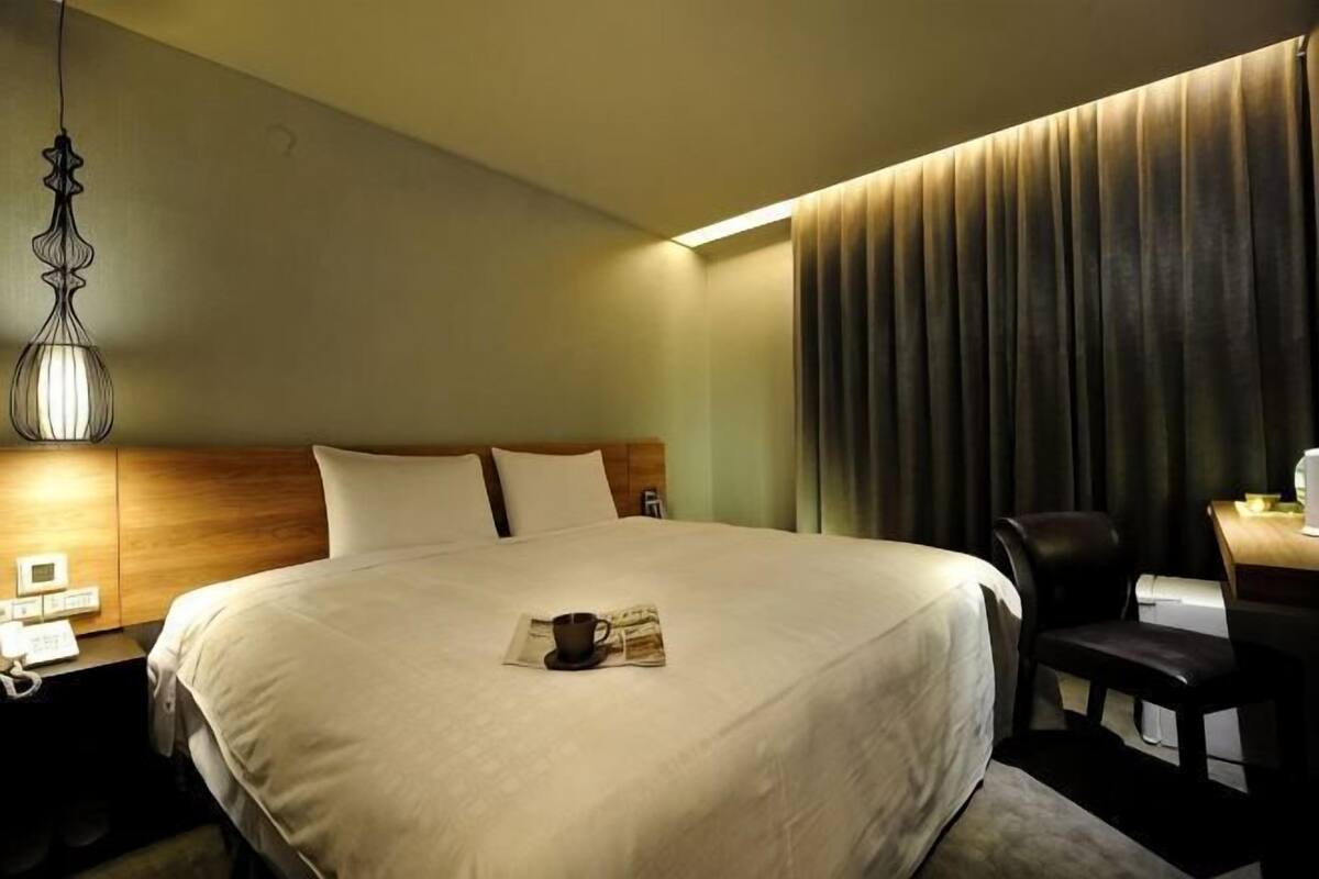 Una habitación de hotel con una cama grande, un escritorio con una silla, una lámpara y una cortina.