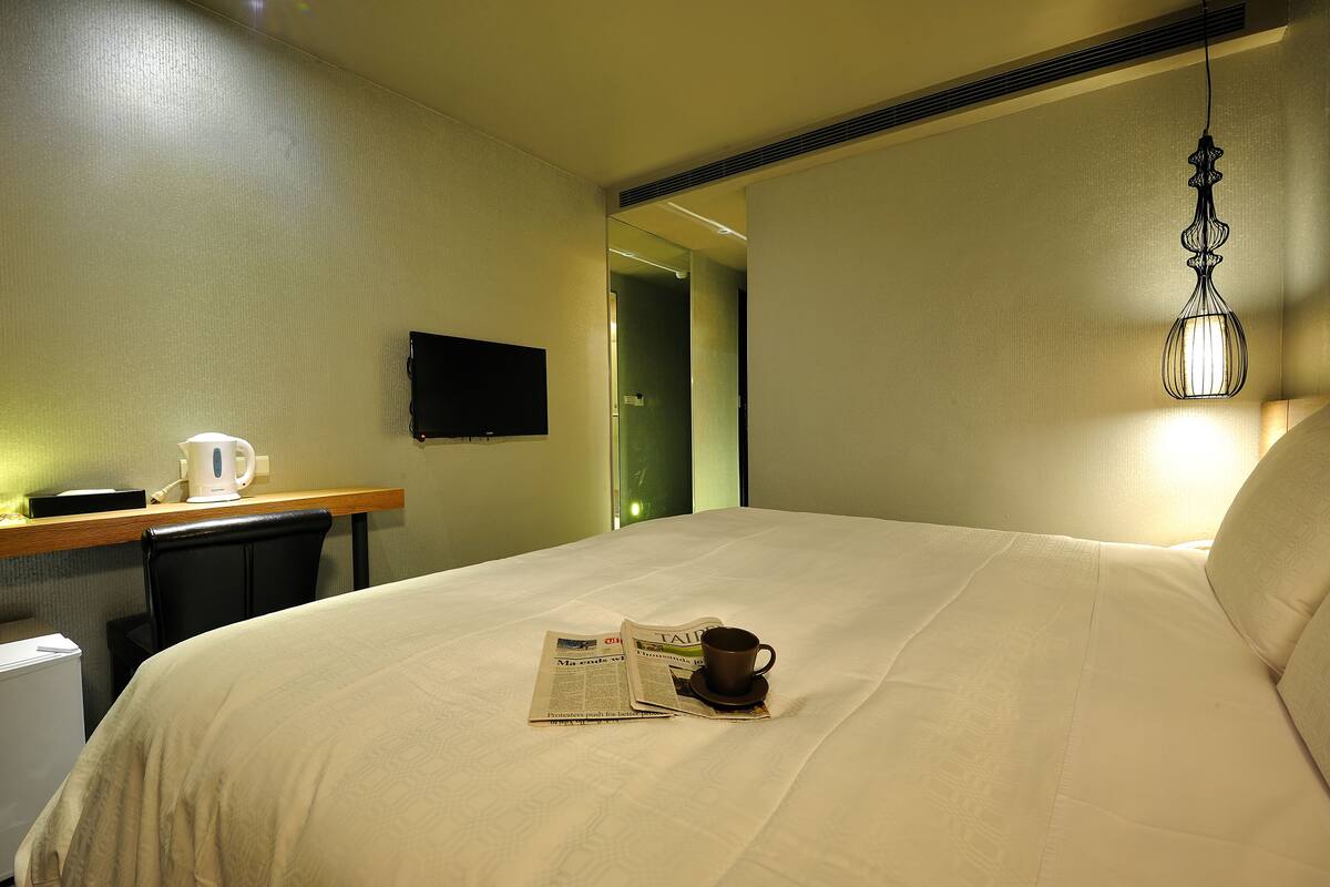 Habitación de hotel con cama, mesita de noche con periódico y taza, televisión de pantalla plana y lámpara colgante.