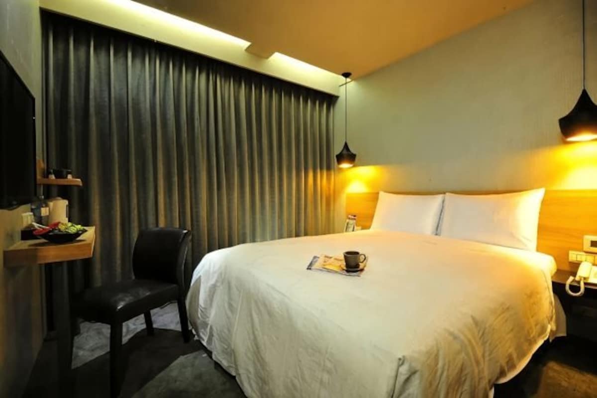 A hotel room with a large bed, a desk with a chair, a lamp, and a curtain.