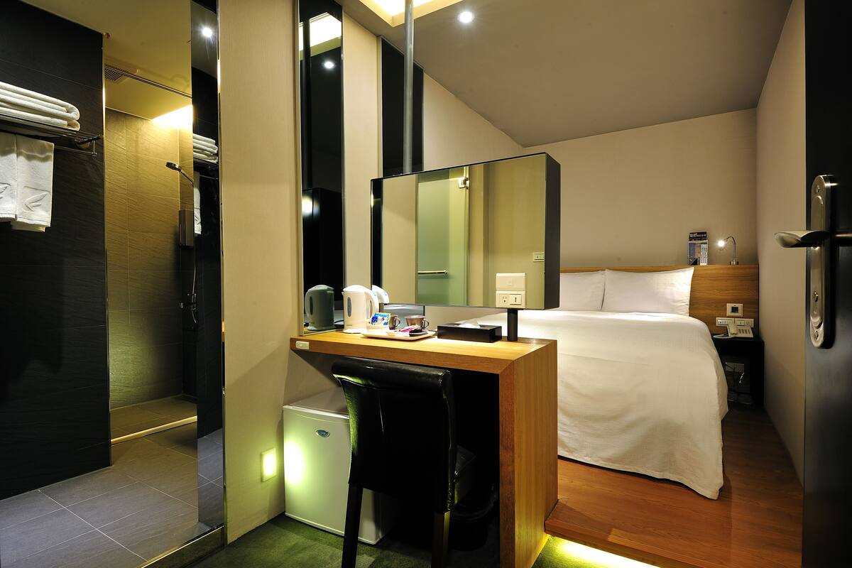 Una habitación de hotel compacta con una cama, un escritorio con silla, un refrigerador pequeño y un espejo.