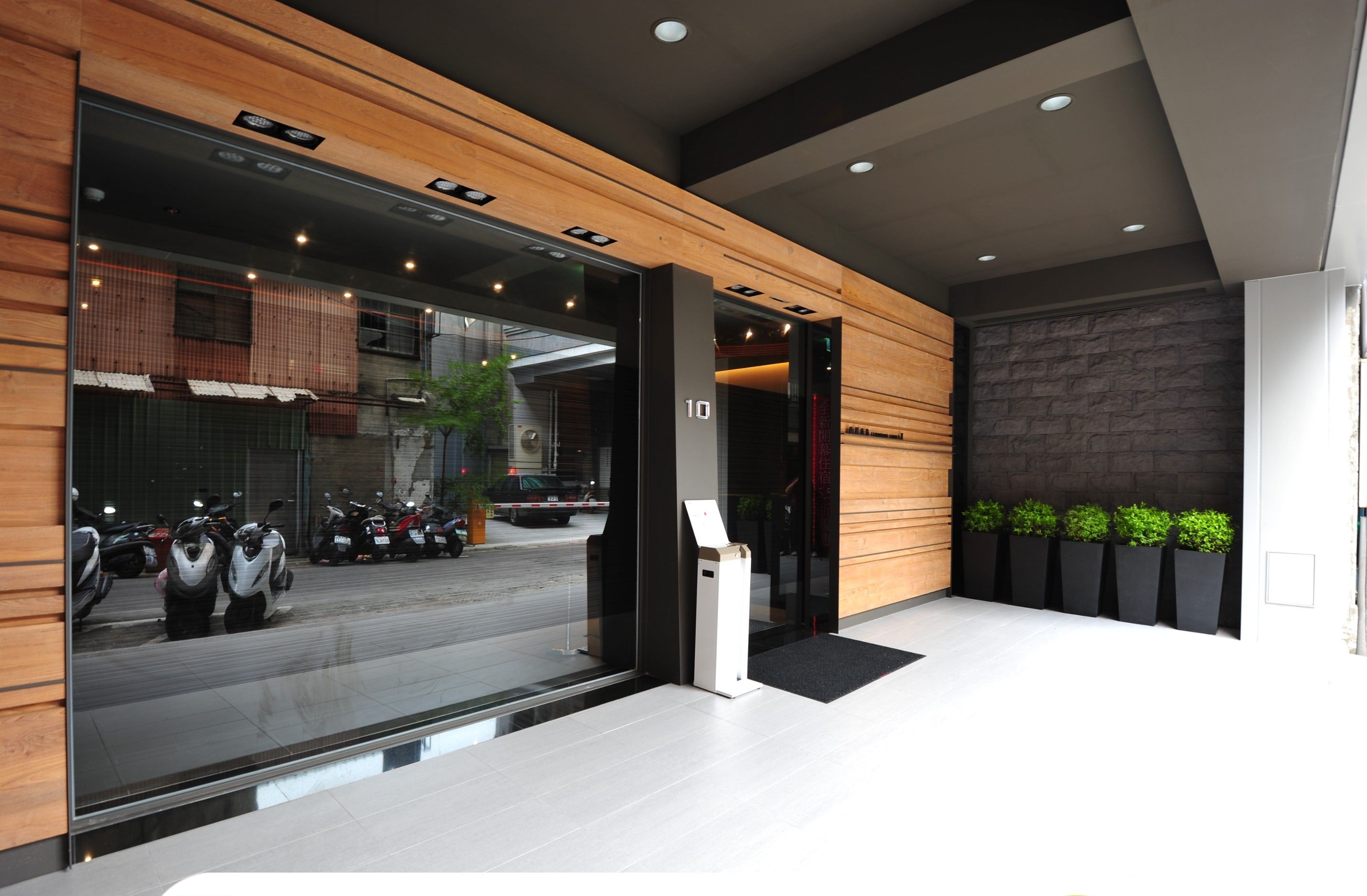 A modern hotel entrance with a registration desk, a view of parked scooters outside, and a stone wall with plants.