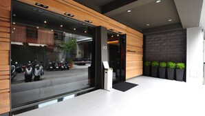 A modern hotel entrance with a registration desk, a view of parked scooters outside, and a stone wall with plants.