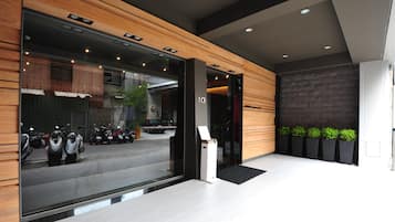 A modern hotel entrance with a registration desk, a view of parked scooters outside, and a stone wall with plants.