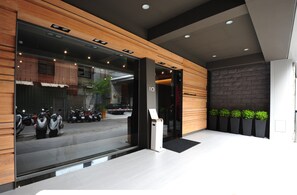 Entrada moderna de hotel com um balcão de recepção, vista de scooters estacionadas e uma parede de pedra com plantas.