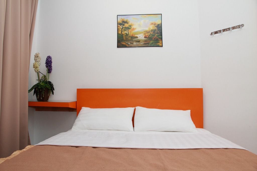 A bedroom with an orange headboard, white bedding, a painting of a landscape, and a potted plant on a nightstand.