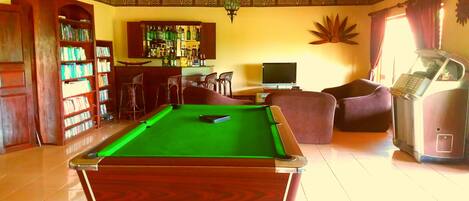 A pool table in a room with a bar area, a television, and a seating area with chairs.