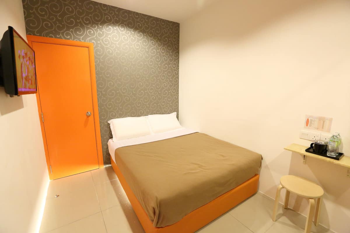 Une chambre d’hôtel avec un lit, un petit tabouret, une étagère avec des articles de toilette et une porte ornée d’un cadre de photo.