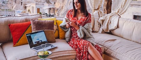A woman sitting on a sofa with a laptop and a tray of food, overlooking a mountainous landscape with traditional architecture.