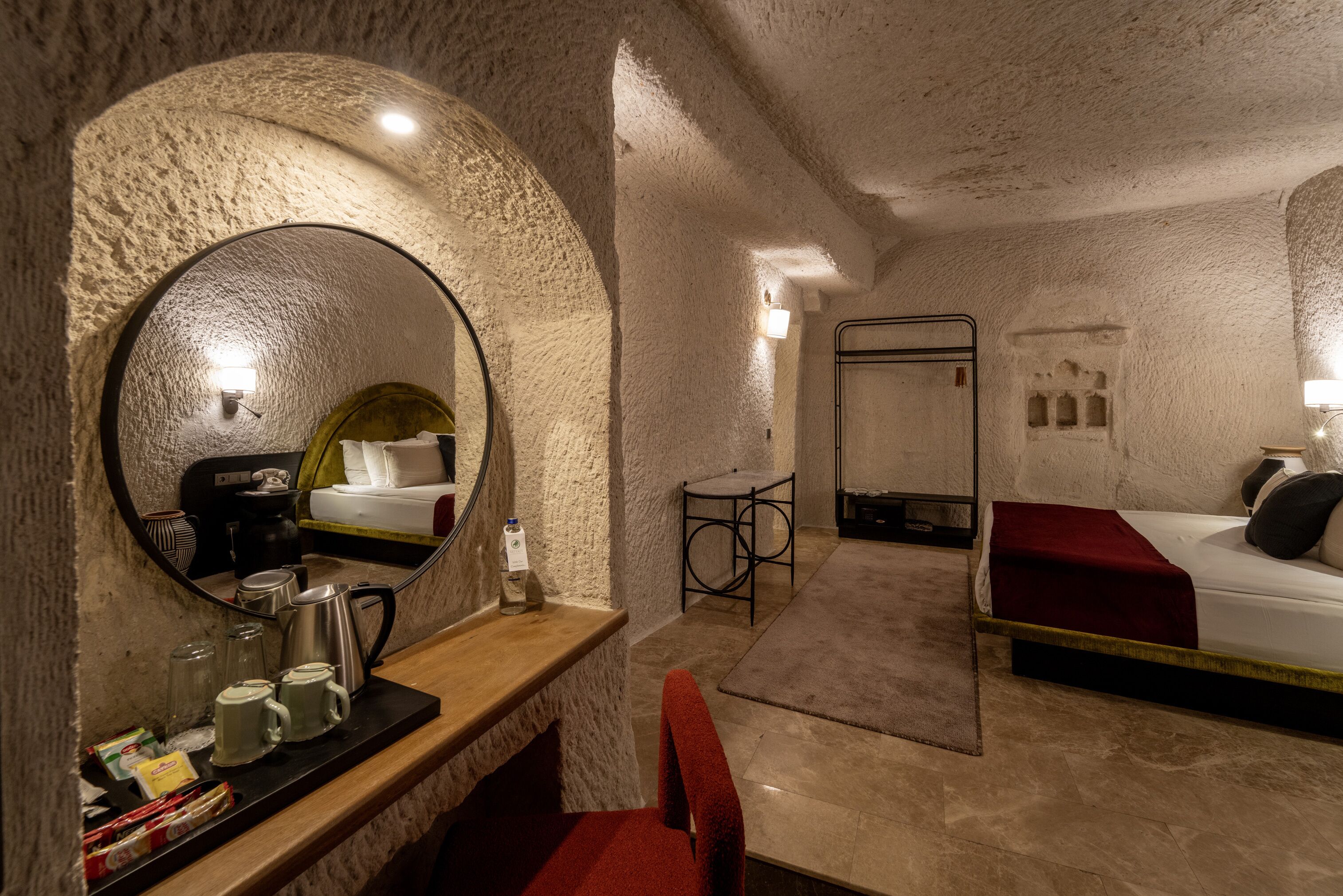 Een knus hotelkamer met een ronde spiegel, een houten bureau met een koffiezetapparaat, een bed met een rode deken en een klein tafeltje met een stoel.