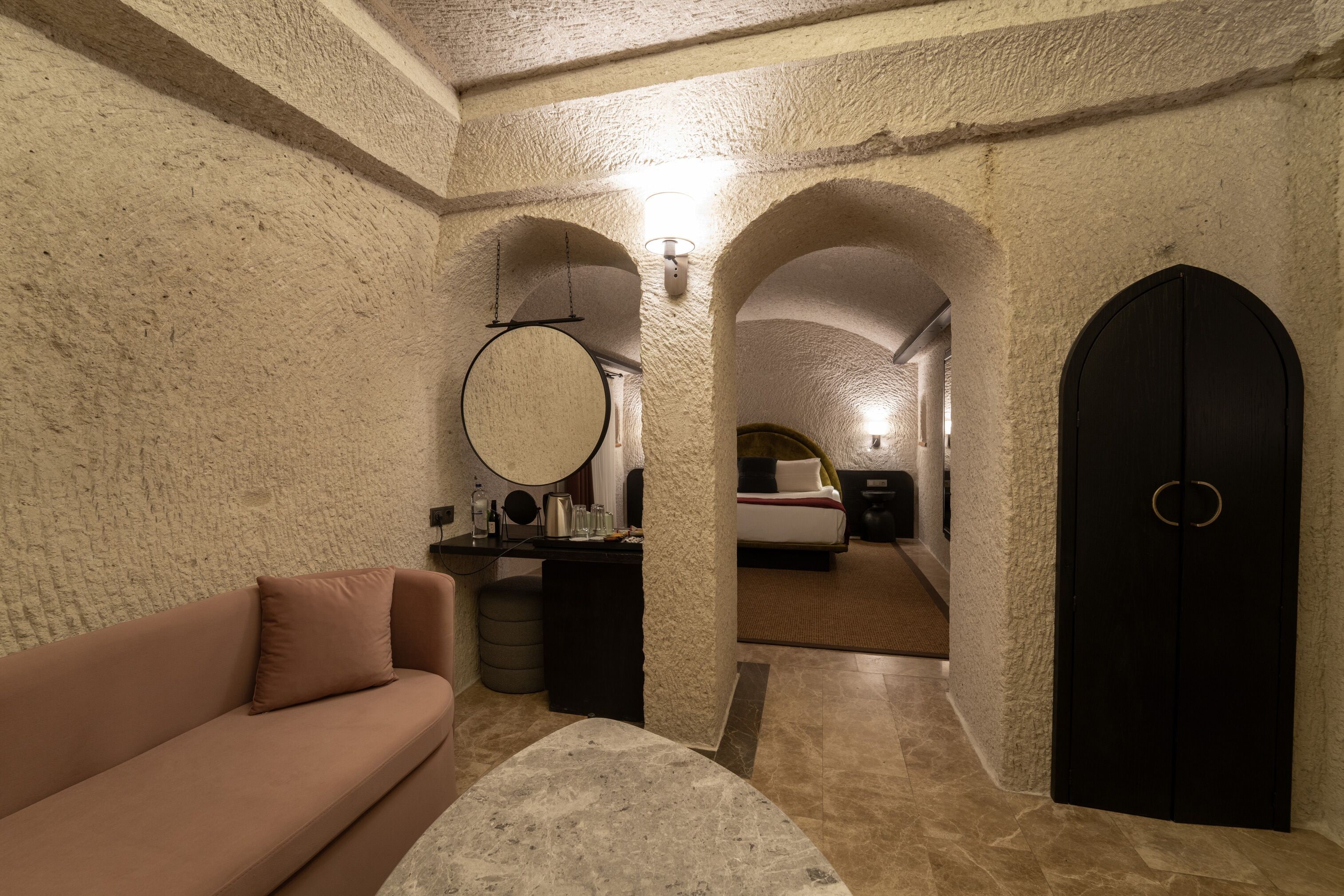 Quarto com parede de pedra, espelho redondo, sofá, cama e porta preta.