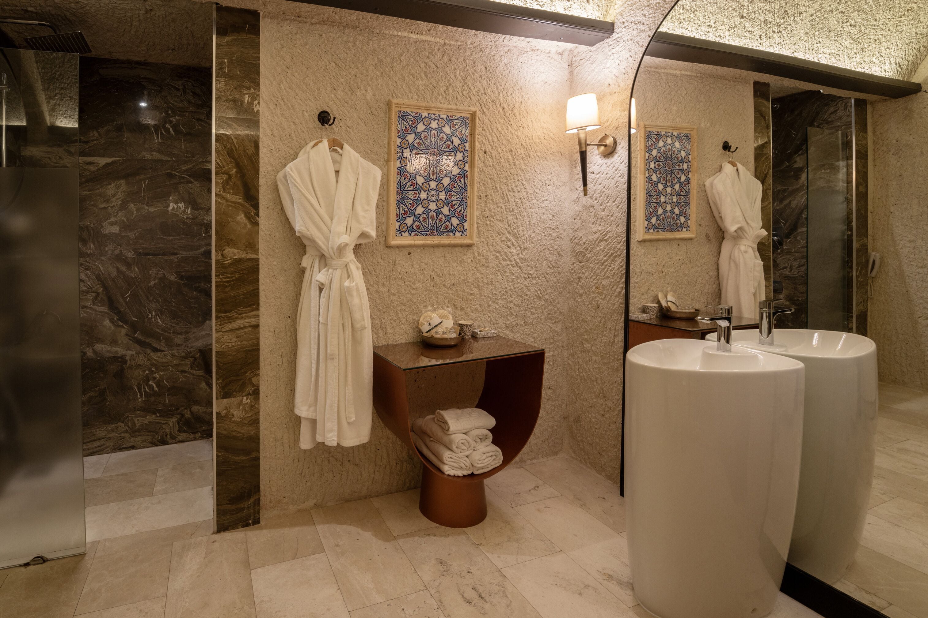 Une salle de bain moderne avec des murs en marbre, un grand miroir, deux vasques et un porte-serviettes avec des peignoirs.