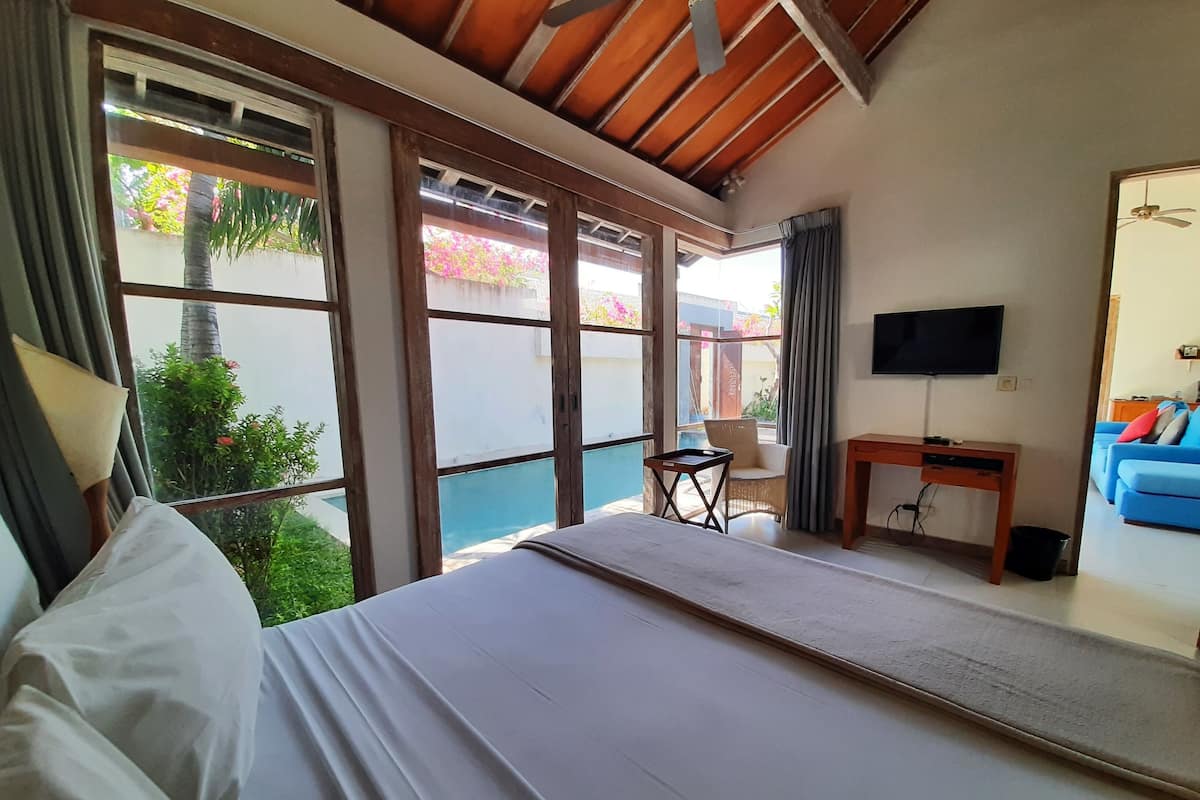 A bedroom with a bed, a desk, a chair, a TV, and a view of a pool and garden.