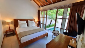 A bedroom with a wooden bed, a nightstand with a lamp, a desk with a computer, and a window with a view of greenery and a pool. - The Decks Bali (Legian)