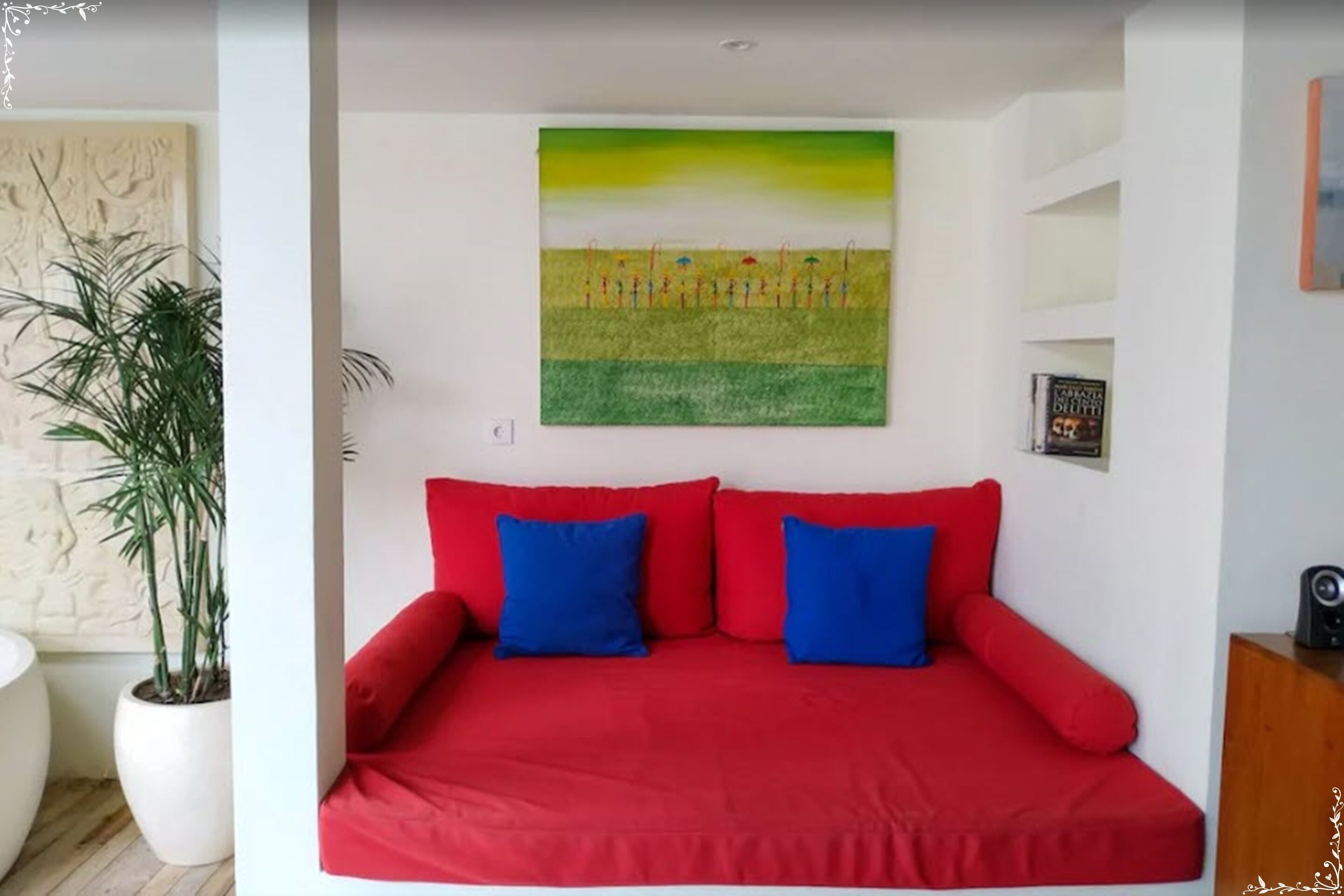 A room with a red sofa bed, a painting of a field, and a potted plant.