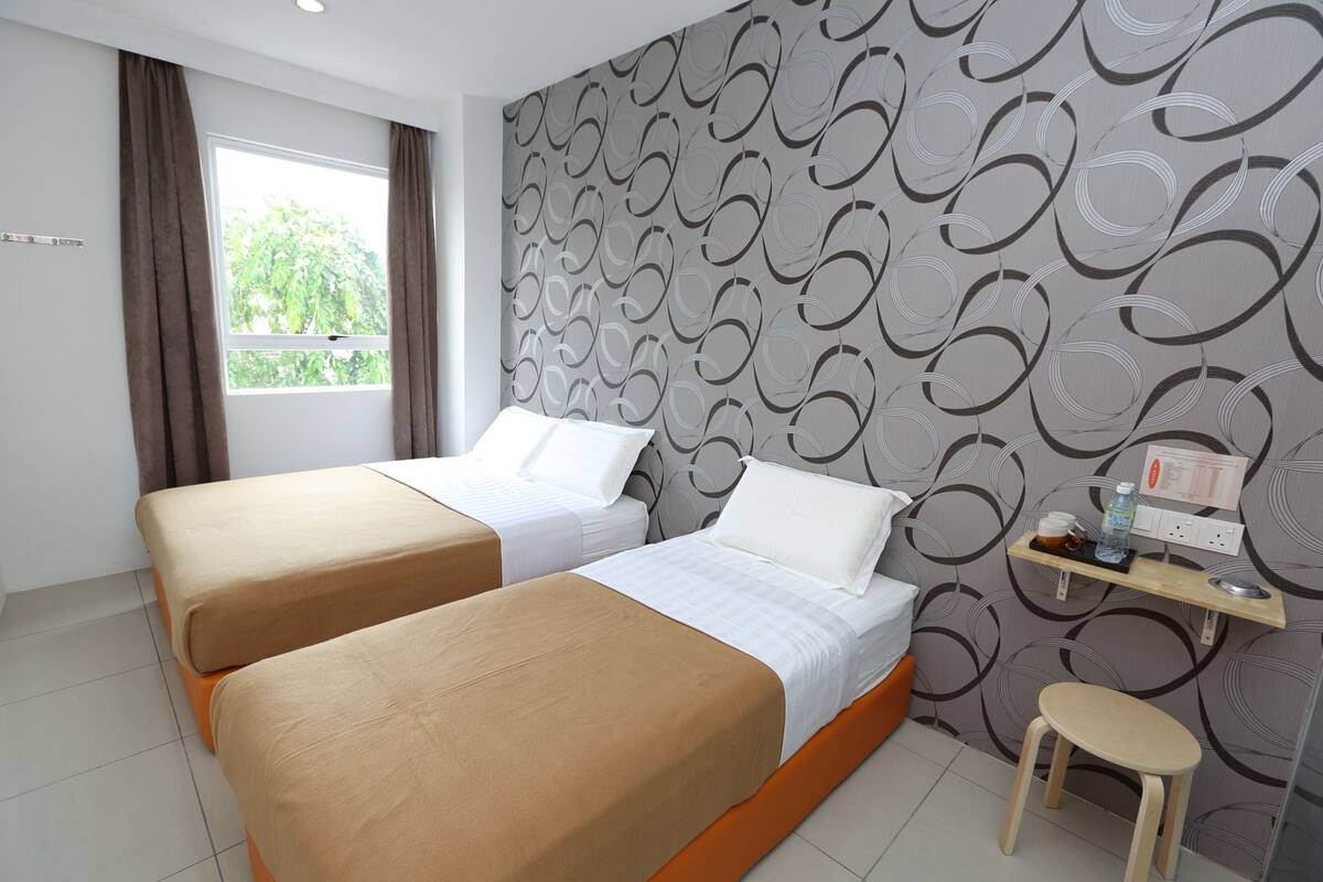 A hotel room with two beds, a window with curtains, a wall with a circular pattern, and a small stool with a shelf above it.