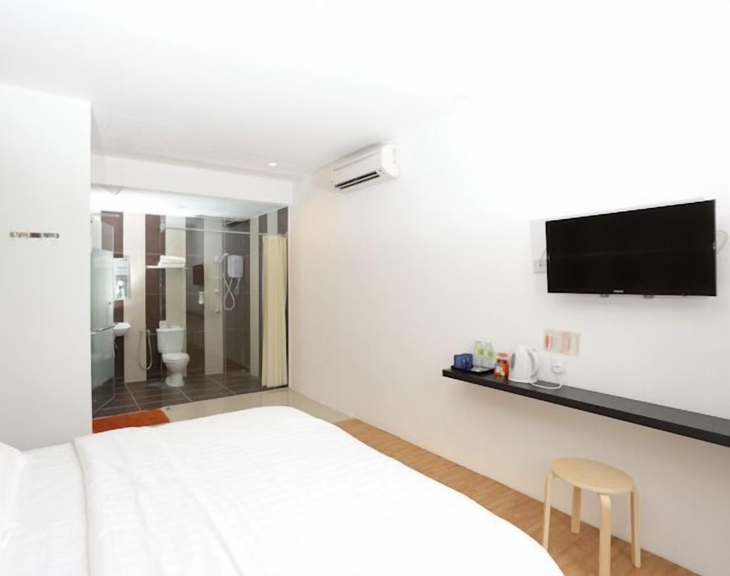 Una habitación de hotel moderna con una cama, un televisor montado en la pared, un taburete pequeño y un baño visible a través de una puerta abierta.