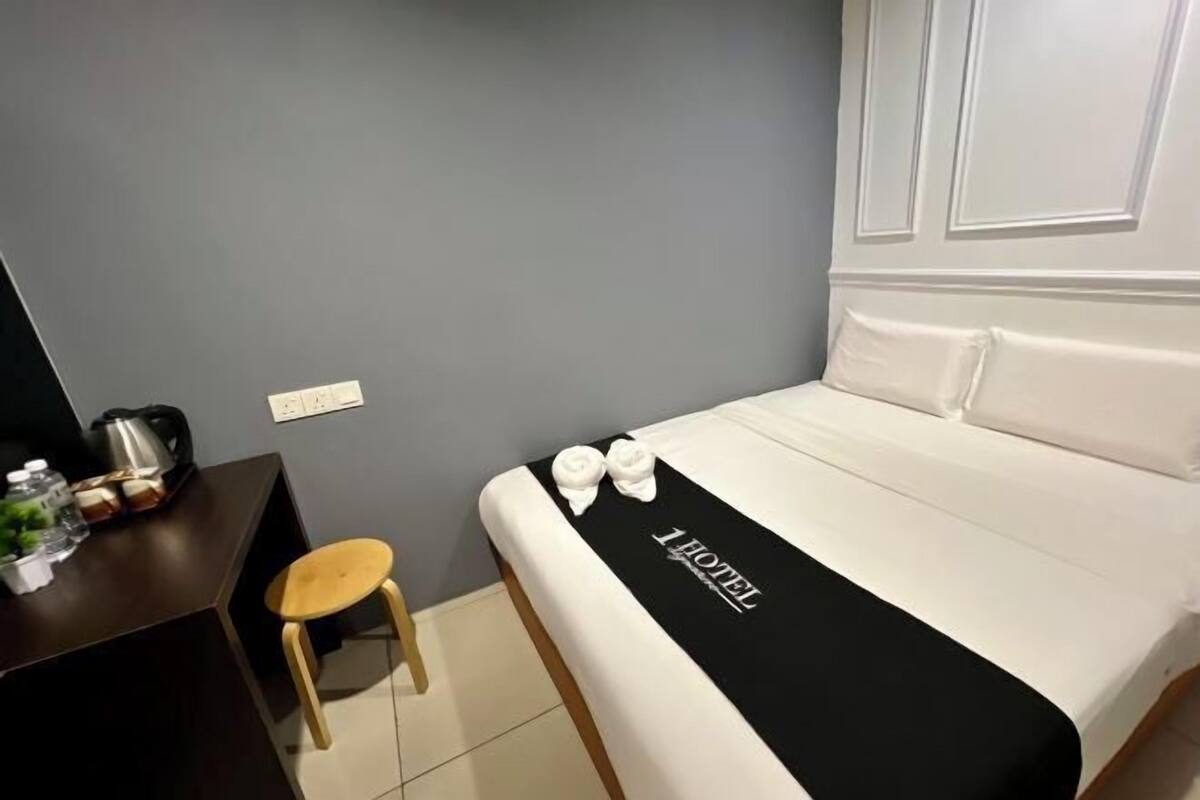 A hotel room with a bed, a desk, a chair, and a small stool.