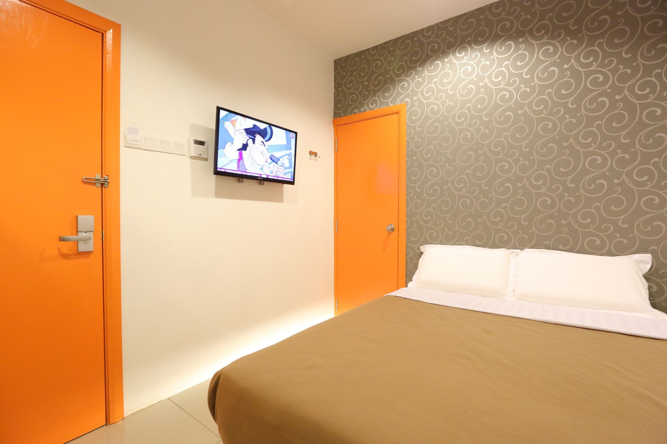 A hotel room with a bed, a television mounted on the wall, and two orange doors.