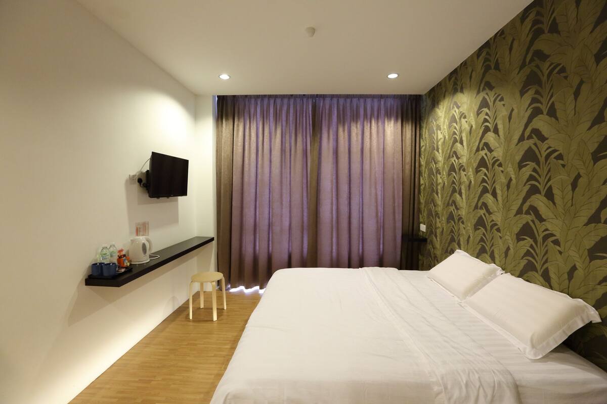 A hotel room with a bed, a TV mounted on the wall, a small table with a chair, and a curtain with a leaf pattern on the wall.